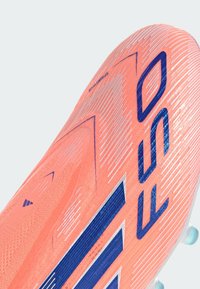 Botín de fútbol naranja con una parte superior texturizada, que presenta acentos azules y un diseño aerodinámico. Suela con tacos para tracción en diversas superficies.