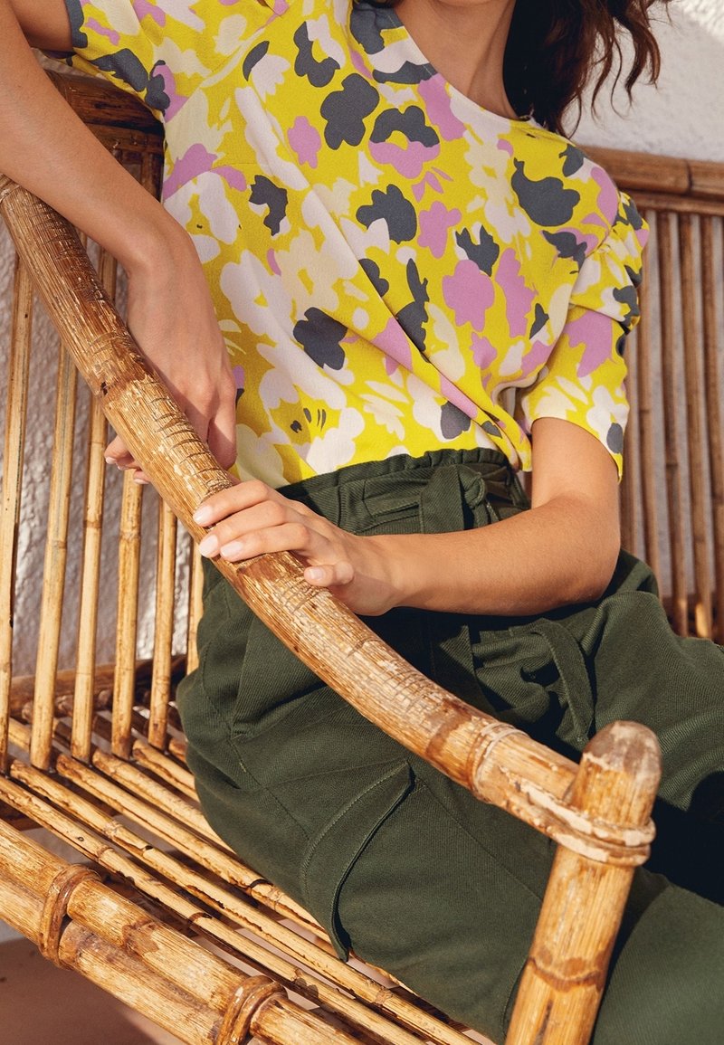 Haut jaune à motifs floraux avec des accents violets, gris et blancs ; pantalon taille haute vert. Les mains reposent sur une chaise en bambou.