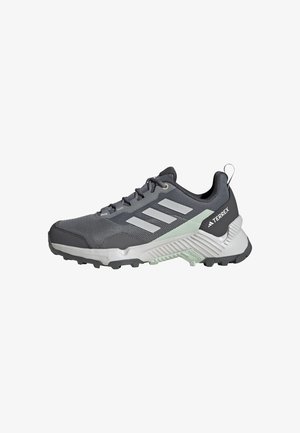 Scarpa sportiva grigia con parte superiore in mesh, tre strisce bianche e suola robusta. Presenta un suola intermedia ammortizzata e una linguetta sul tallone.