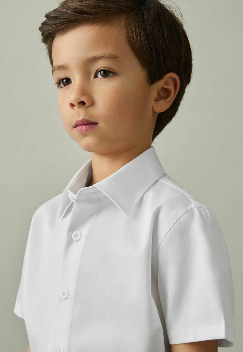Jeune garçon aux cheveux courts et foncés, portant une chemise blanche à manches courtes boutonnée, regardant légèrement sur le côté sur un fond uni.