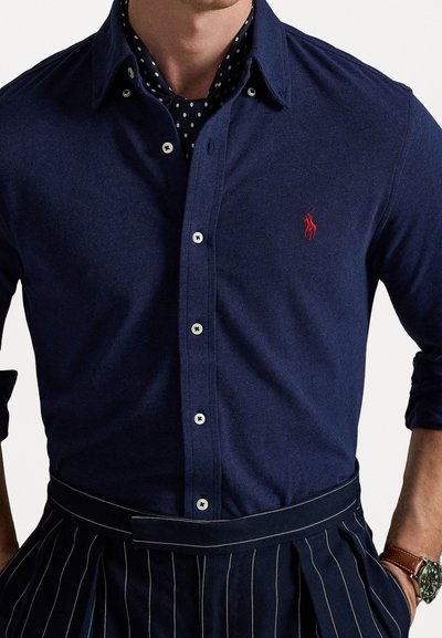 Mornarji modra srajca na gumb z rahlo teksturo, z belimi gumbi in rdečim logotipom. Oblečena čez srajco z vzorcem pik in kombinirana z črtastimi hlačami.