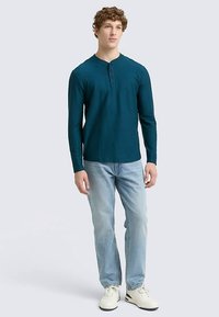 Daljša majica henley v barvi modrozelene s trirazredno zapenjanjem na gumbe, kombinirana z svetlomodrimi kavbojkami in belimi supergami, prikazana na nevtralni podlagi.