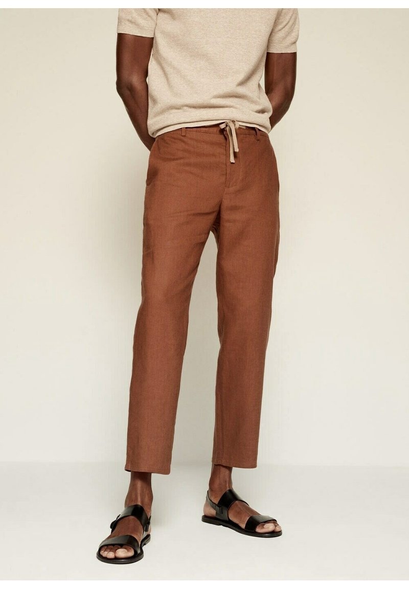 Pantalons en lin marron avec une coupe droite, taille élastique avec cordon de serrage, et poches latérales, associés à des sandales noires. Texturé lisse et design léger.