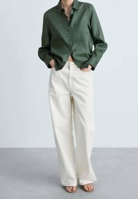 Camicia verde in lino con bottoni abbinata a pantaloni larghi color crema e sandali marrone chiaro, creando un look rilassato e informale.