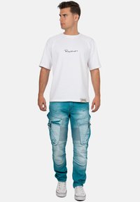 T-shirt blanc avec texte noir, associé à un pantalon cargo bleu clair doté de poches et d'accents texturés, porté avec des baskets blanches.