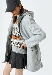 Cappotto grigio con cappuccio e finitura testurizzata, indossato sopra un maglione con motivo caratterizzato da tonalità calde, abbinato a una gonna plissettata di grigio scuro.