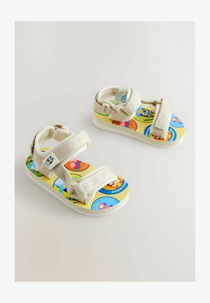 Vaikų sandalai su šviesiai beige audiniu, reguliuojamomis dirželiais ir spalvinga, raštuota padi, vaizduojančia animacinių filmų personažus.