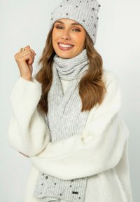 Grijze gebreide beanie met decoratieve kralen, bijpassende geribbelde sjaal en een witte oversized trui. Zachte textuur, warme uitstraling en functioneel ontwerp.