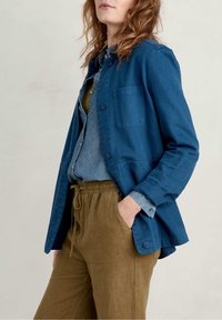 Žena oblečená v modré saková bundě na knoflíky přes denimovou košili a hnědých kalhotách se stahovacími šňůrkami, s rukou v kapse, stojí proti jednobarevnému pozadí.