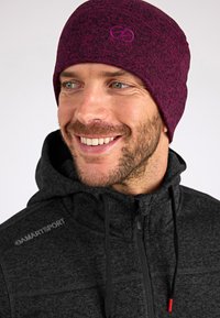 Gorro de punto burdeos con textura, que presenta un logotipo en el frente. Se lleva con una chaqueta oscura con cremallera que muestra costuras visibles y cordones ajustables.