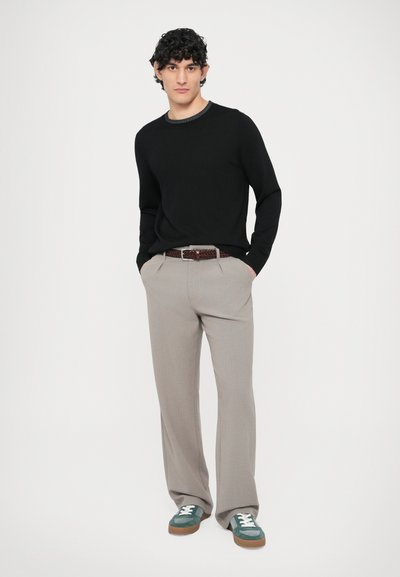 Czarny sweter, beżowe plisowane spodnie, brązowy pleciony pasek, zielone zamszowe trampki z białymi sznurówkami, na jednolitym białym tle.