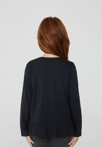 Námořnická modrá halenka s dlouhým rukávem a volánkem, vyrobená z měkkého materiálu. Jednoduchý design, kulatý výstřih a bez viditelných vzorů či kovových prvků.