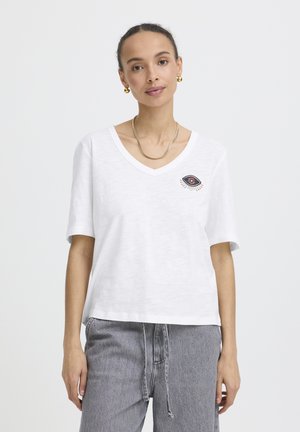 Femeie purtând un tricou alb cu decolteu în V, decorat cu un design de ochi brodat, și blugi gri cu talie înaltă, accesorizată cu cercei și colier aurii.