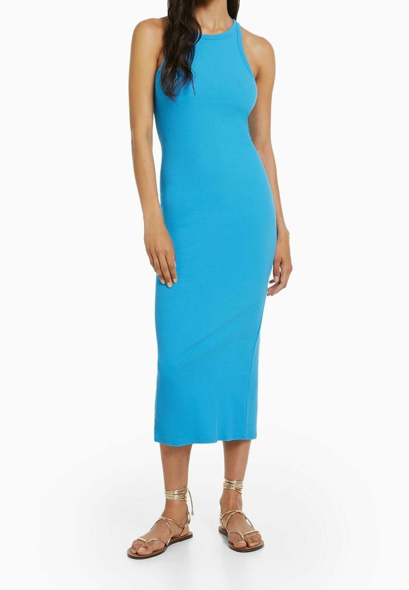 Femme portant une robe midi sans manches, côtelée, de couleur bleu vif, et des sandales plates à lanières dorées, debout devant un fond blanc.