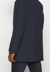 Námořnicky modrá parka bunda s hladkou texturou, dlouhými rukávy a černou logo nášivkou na levém rameni, doplněná tmavými džínami na spodním okraji.