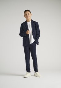 Granatowy garnitur z dopasowaną marynarką i spodniami, w połączeniu z białym podkoszulkiem i białymi trampkami, wykonany z gładkiego materiału i w dopasowanym kroju.