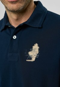 Mornarska polo majica s tri gumba i teksturiranom tkaninom. Dekorativna bež grafika na prsima s konturama nalik karti.