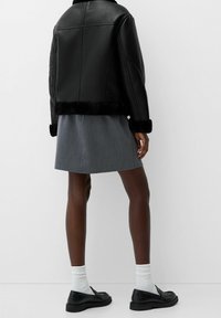 Zwarte imitatieleren jas met een bontvoering bij de kraag en manchetten, gecombineerd met een grijze rok. Zwarte loafers en witte sokken maken de outfit af.