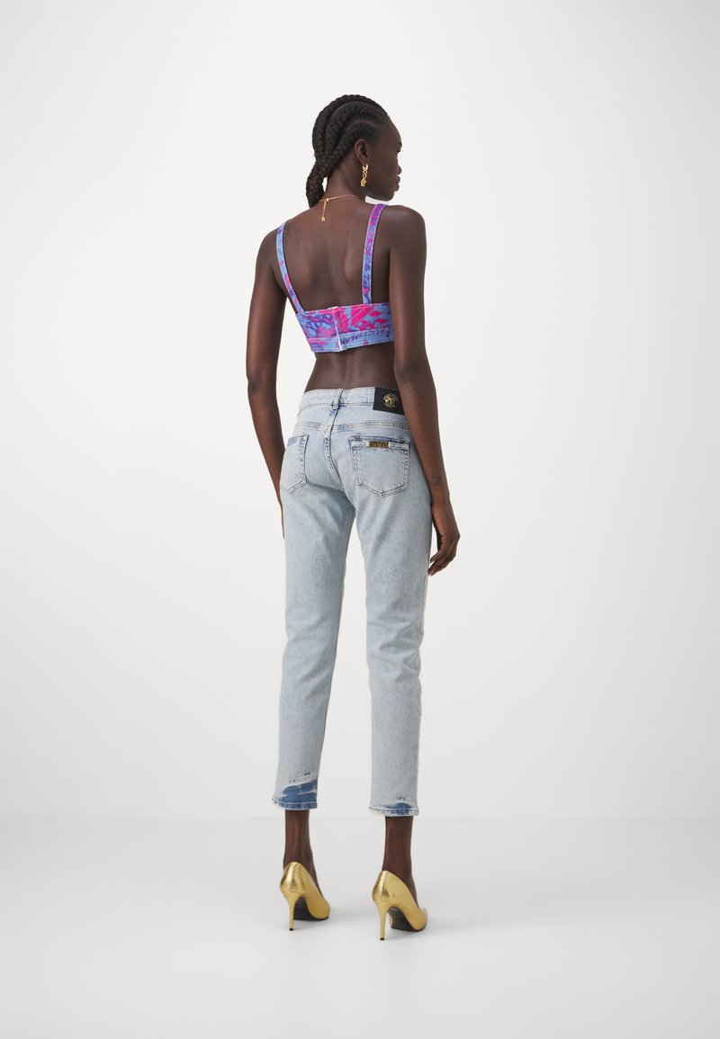 Kobieta z warkoczem, ubrana w różowo-niebieski crop top, jasnoniebieskie dżinsy z wystrzępionymi brzegami i złote szpilki, zwrócona plecami na białym tle.