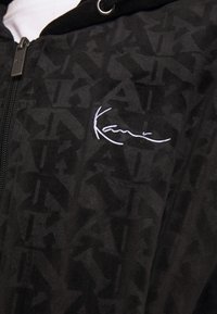 Felpa nera con zip, caratterizzata da un motivo testurizzato e un logo ricamato bianco sul petto. Il tessuto ha una finitura liscia e una sensazione morbida al tatto.