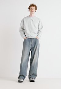 Sweat-shirt gris clair avec des poignets côtelés, associé à un jean bleu ample. La mannequin porte un collier simple. Fond minimaliste.