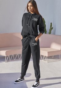 Schwarzer Hoodie mit dem Text "GENIESSE JEDEN MOMENT DEINES LEBENS", kombiniert mit passenden Jogginghosen und schwarzen Sneakers. Weicher Stoff, lässige Passform, Kordelzugdetails.