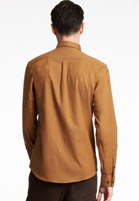 Camicia marrone a maniche lunghe con una texture liscia, polsini con bottoni e una piega sul retro. Presenta un colletto classico e un orlo leggermente curvato.