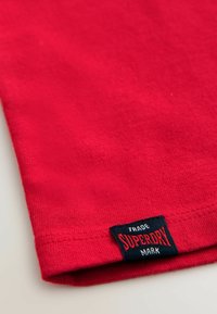 Červené bavlněné tričko s hladkou texturou a skládaným lemem. Na černé etiketě je nápis "SUPERDRY" v bílé a červené barvě.
