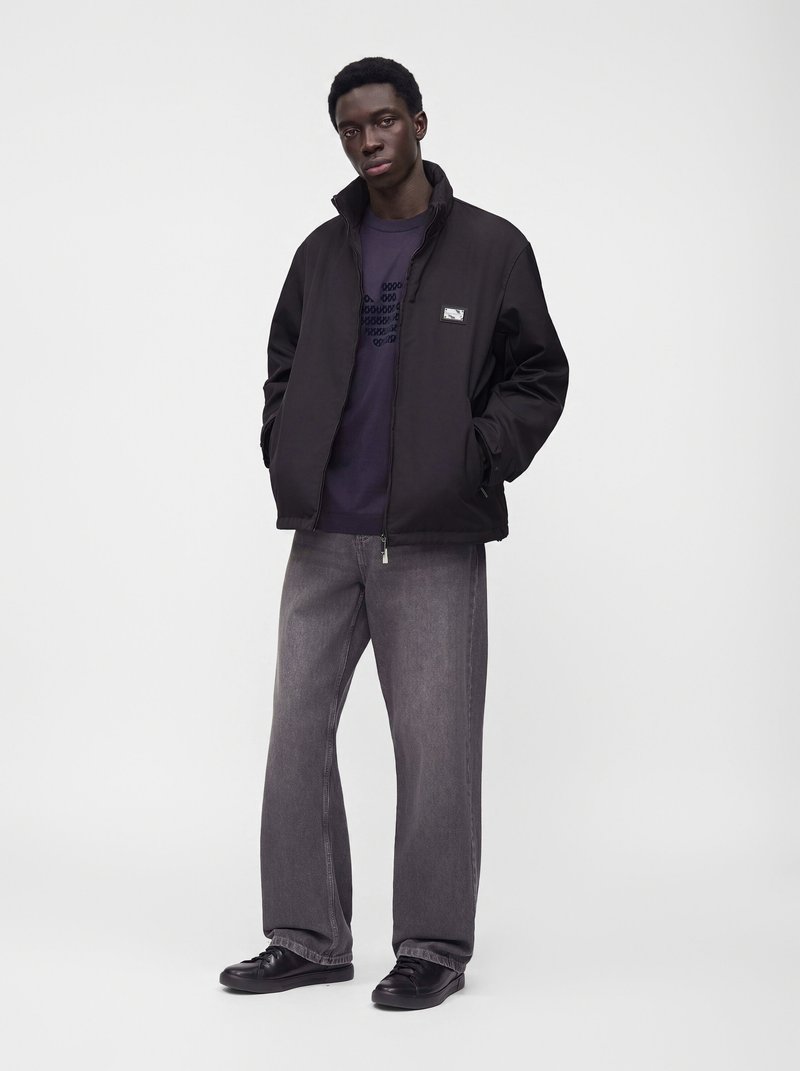 Jeune homme debout, mains dans les poches, portant une veste noire, une chemise bleu foncé, un jean gris et des chaussures noires sur un fond blanc uni.
