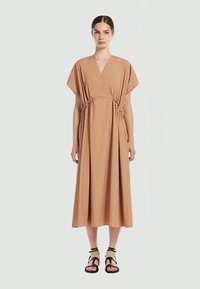 Femeie îmbrăcată cu o rochie midi bej, cu mâneci scurte și talie strânsă, purtând sandale negre cu barete, stând pe un fundal simplu.