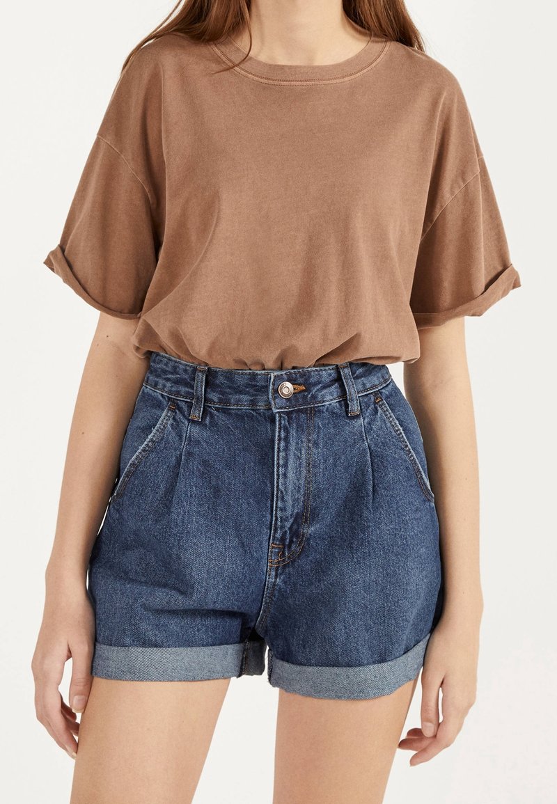 Femme portant un t-shirt marron à manches courtes rentré dans un short en jean bleu à taille haute et revers, sur un fond uni.