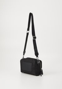 Sac bandoulière en tissu noir de forme rectangulaire, avec une poche avant, des accessoires en métal argenté et une sangle réglable accentuée par un logo.