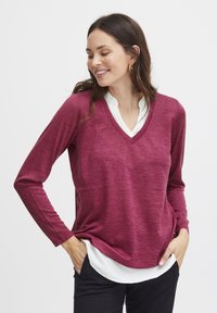 Maglietta bordeaux a maniche lunghe con scollo a V e finitura testurizzata, sovrapposta a una camicia bianca con colletto; vestibilità rilassata con orlo curvato.