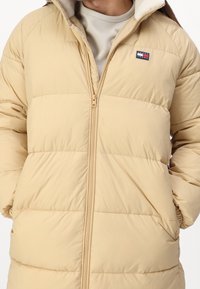 Puffet beige jakke med lynlås foran, der har en blød indvendig krave, vandret syninger og et lille logo på brystet.