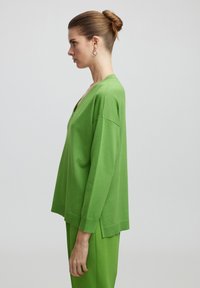 V-Ausschnitt, langärmliger Pullover aus leuchtend grünem Strickstoff; lockere Passform mit seitlichen Schlitzen. Kombiniert mit passenden grünen Hosen.