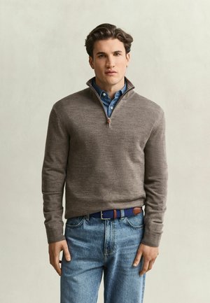 Young man wearing a gray quarter-zip sweater over a blue button-up shirt, blue jeans, and a blue woven belt, standing against a plain background.