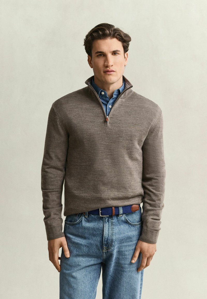Jeune homme portant un pull gris à fermeture éclair quart sur une chemise bleue à boutons, un jean bleu et une ceinture tressée bleue, debout devant un fond uni.