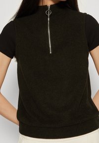 Dunkelgrünes ärmelloses Oberteil mit hohem Kragen, silbernem Reißverschluss und gerippter Textur, über einem schwarzen kurzärmeligen Shirt getragen.