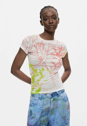 Kvinde med fletninger, iført en hvid t-shirt med korte ærmer med røde og grønne blomsterprint og blå mønstrede jeans, står med hænderne bag ryggen.