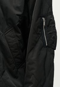 Μανίκι μαύρου καπιτονέ μπουφάν με εμφανές ασημί φερμουάρ σε τσέπη και ραφές σε ματ ύφασμα.