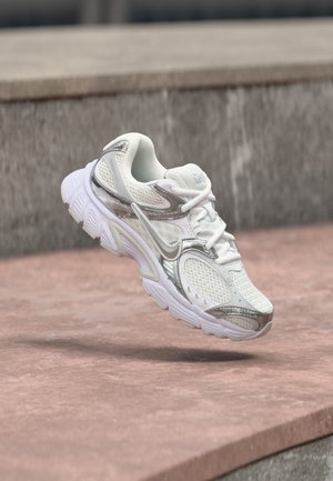 Scarpa da corsa in mesh bianca e argento che fluttua sopra una sporgenza in cemento rosa con uno sfondo di muro in pietra grigia.