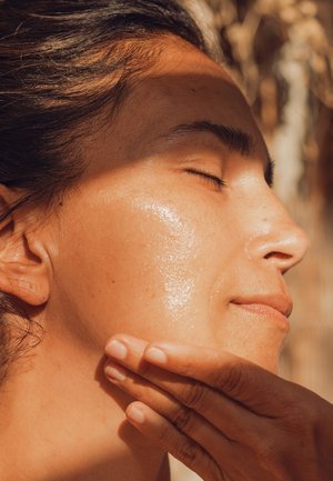 Close-up of a face with sun-kissed skin, smooth texture, and a glossy sheen. Hand gently touching cheek. Hair is dark and wavy.