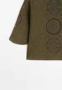Olive green fabric with laser-cut and embroidered geometric floral patterns on the sleeve and front panel of a garment.