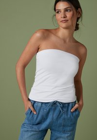 Top blanc sans bretelles en tissu doux, au design ajusté, associé à un pantalon en denim bleu décontracté avec taille à cordon et poches latérales sur fond vert.