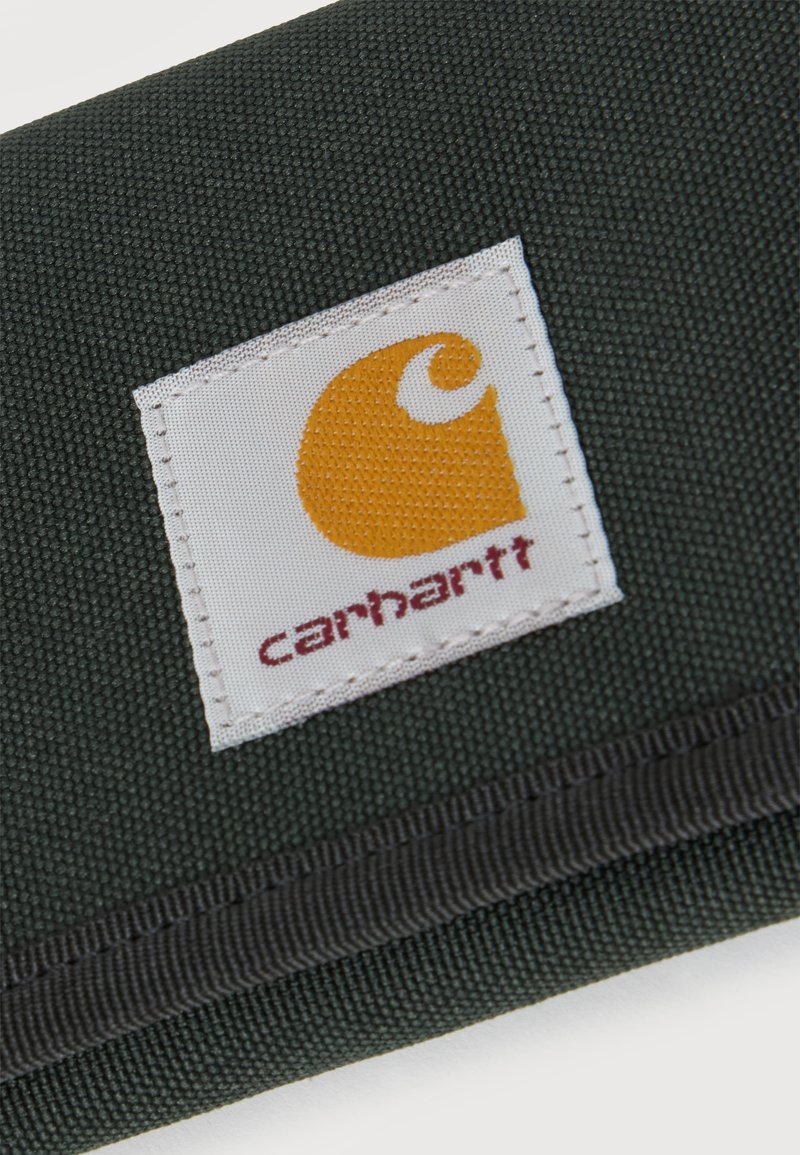 Tela verde oscura con una etiqueta cuadrada blanca cosida que muestra el logo naranja de Carhartt y texto rojo.