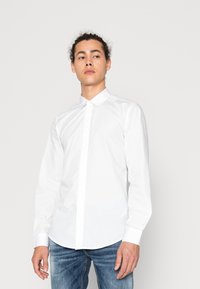 Witte lange mouwen blouse met een spitse kraag, knoopsluiting aan de voorkant en een gebogen zoom. Gemaakt van gladde stof, gedragen met blauwe jeans.