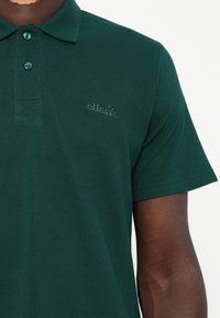 Polo de cor verde escura, feito de tecido texturizado, com mata-borrão de três botões e um discreto logo em relevo no peito.