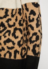 Gebreide trui met een tan lichaam voorzien van zwarte luipaardprintpatronen, afgewerkt met een effen zwarte onderband. Zacht, gestructureerd materiaal.