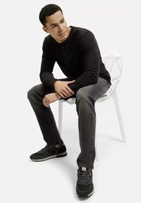 Czarna koszulka z długim rękawem, szare dżinsy i czarne buty sportowe. Siedzi na białym krześle o minimalistycznym designie.