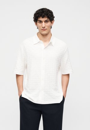 Jeune homme aux cheveux bouclés foncés portant une chemise blanche texturée à manches courtes boutonnée et un pantalon noir, mains dans les poches, fond neutre.